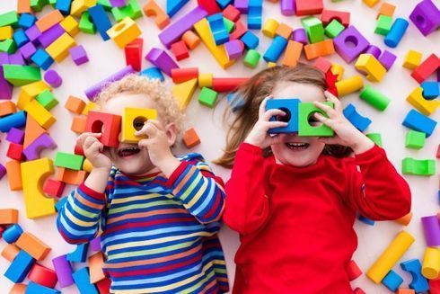 Zwei lachende Kinder halten bunte Bauklötze vor ihre Augen, umgeben von weiteren Bauklötzen.