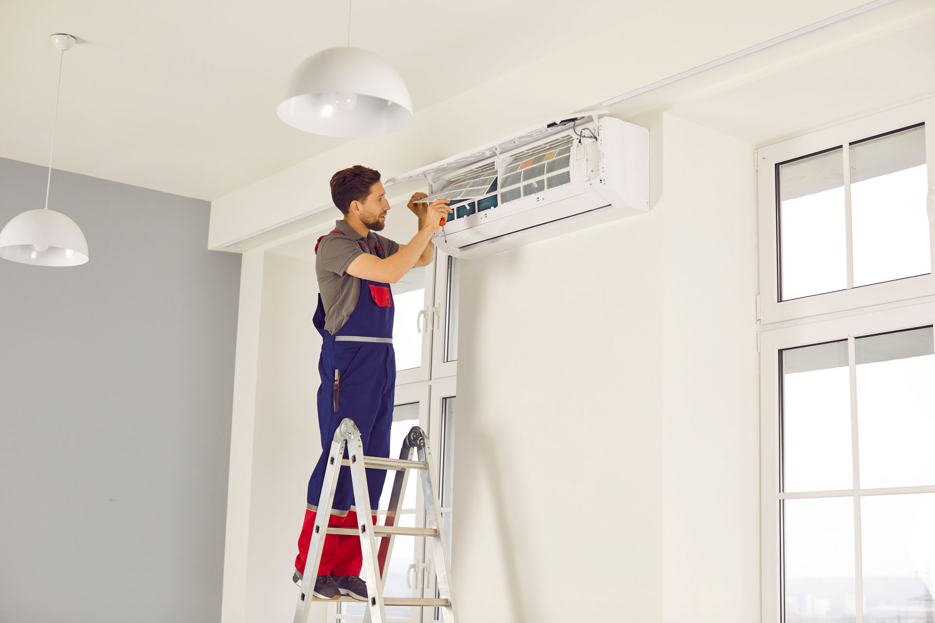 Man in overalls on ladder, fixing an air conditioner indoors. Maintenance
