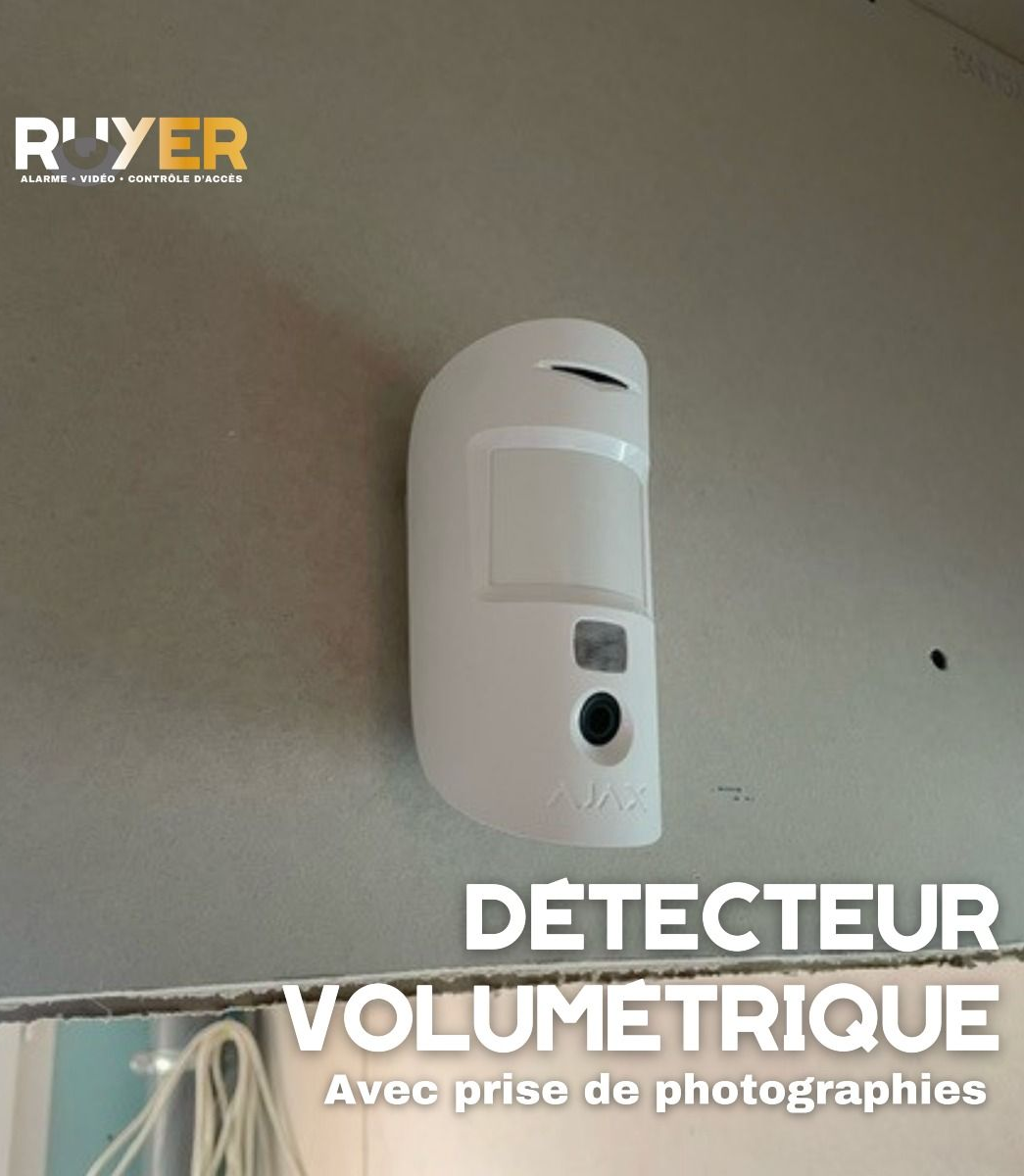 Détecteur volumétrique blanc fixé sur un mur gris. Inscription : « Détecteur volumétrique ».