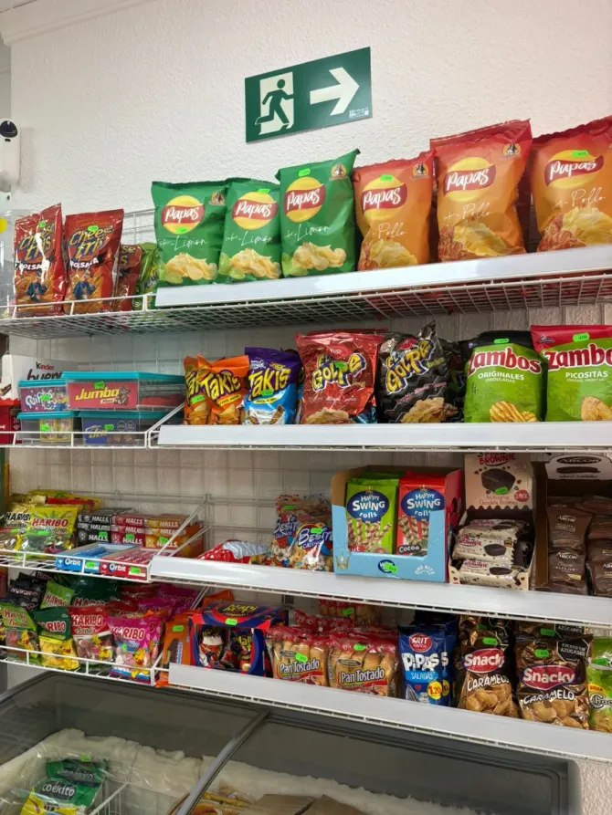 Estanterías de una tienda de conveniencia repletas de coloridas patatas fritas y caramelos, con un letrero de salida encima.