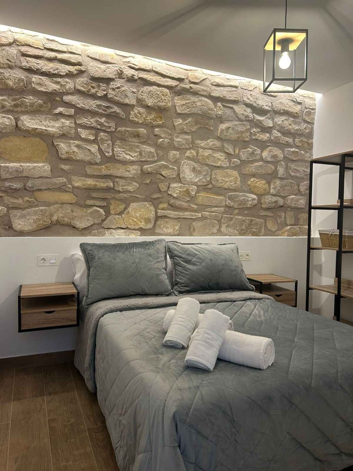 Un dormitorio en tonos grises con una pared decorativa de piedra, una cama con ropa de cama gris y toallas dobladas, y una mesita de noche flotante.