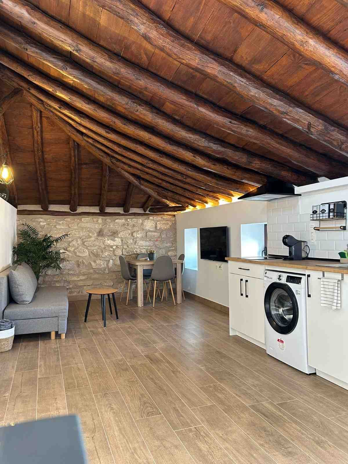 Un apartamento tipo loft de planta abierta con techo rústico con vigas de madera, pared de piedra, cocina americana y sofá gris.