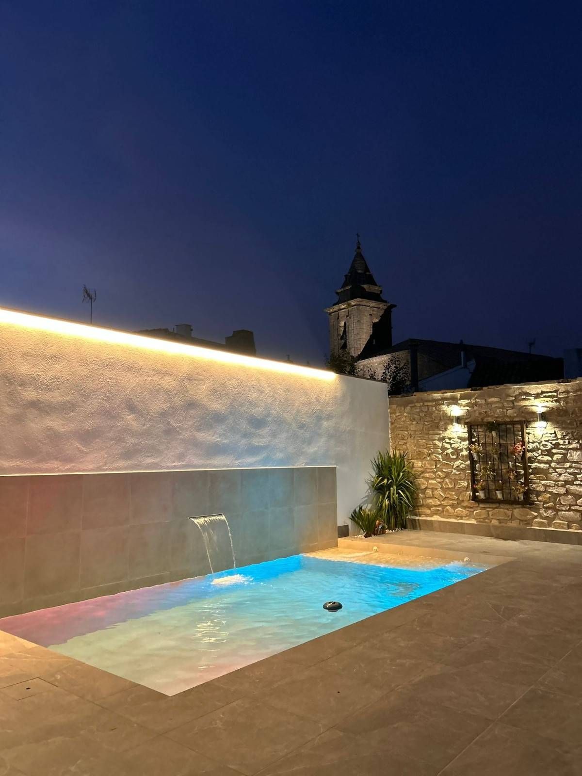 Una pequeña piscina iluminada con una fuente de agua por la noche, situada junto a un muro de piedra, con el campanario de una iglesia al fondo.