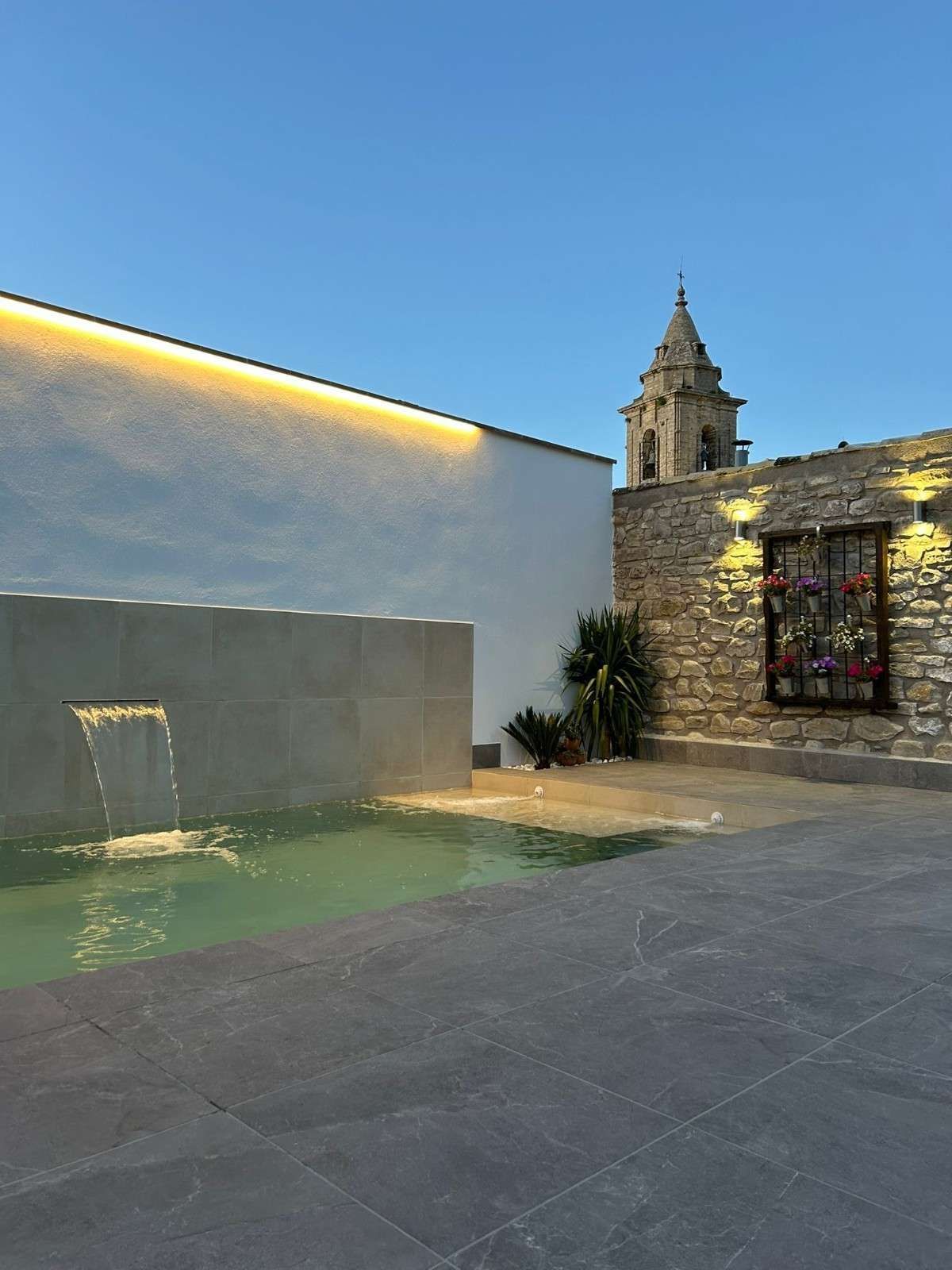 Patio moderno con muro de piedra, piscina rectangular con fuente y vistas a un campanario de piedra.