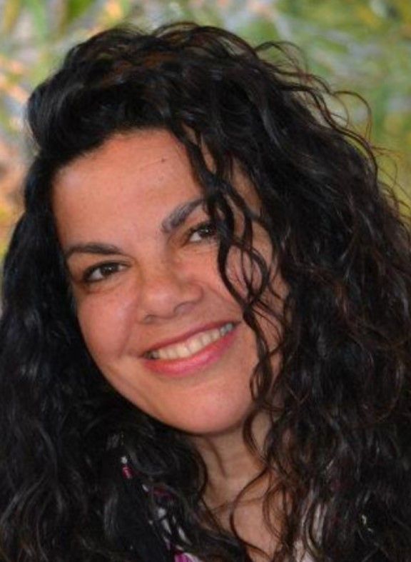 Una mujer con cabello oscuro, largo y rizado sonríe y mira hacia la cámara.