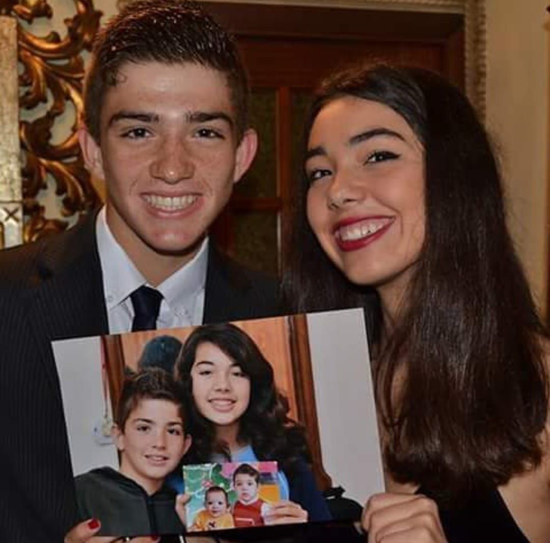 Un hombre joven y una mujer sonriendo, sosteniendo una foto de ellos mismos cuando eran niños.