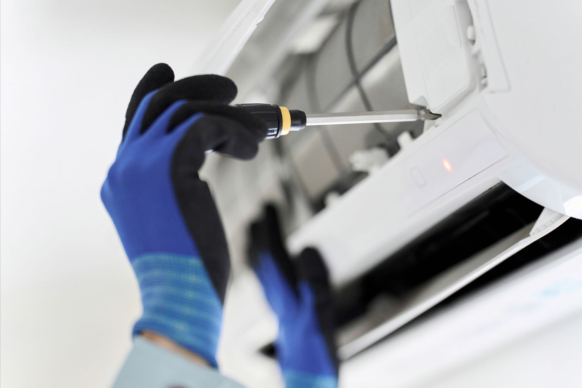 Des mains gantées de bleus manipulent un tournevis sur un climatiseur blanc.