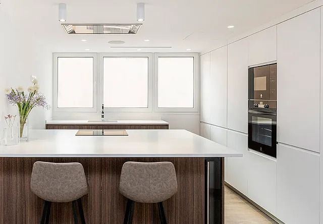 Una cocina con gabinetes blancos, una isla grande, dos taburetes y una ventana.