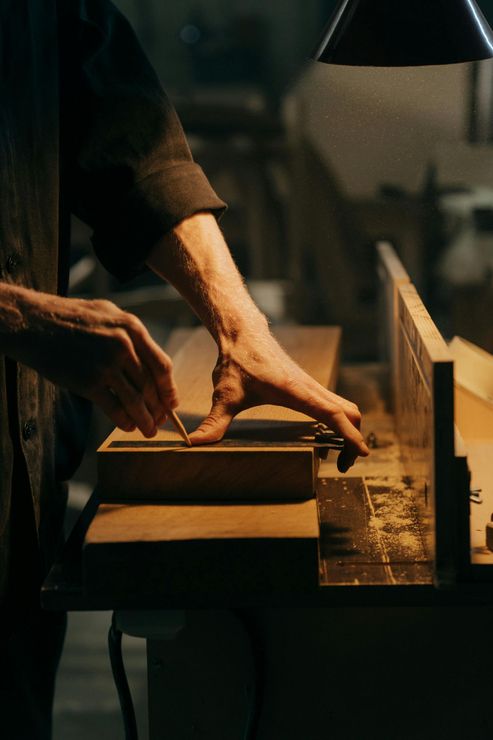 Manos de un carpintero dando forma a la madera con una herramienta en un banco de trabajo, iluminado por una lámpara.