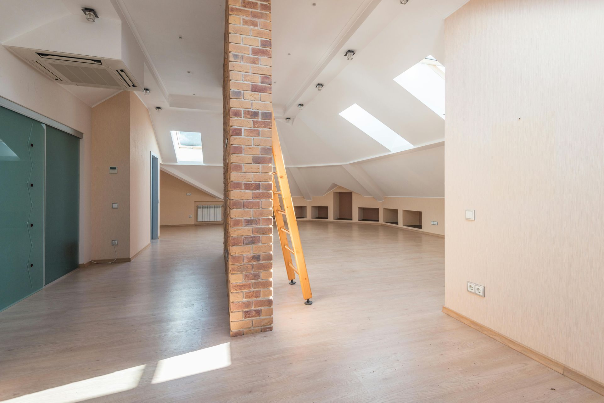 Amplio ático con columna de ladrillo, claraboyas y escalera de madera. Paredes beige y suelos de madera clara.