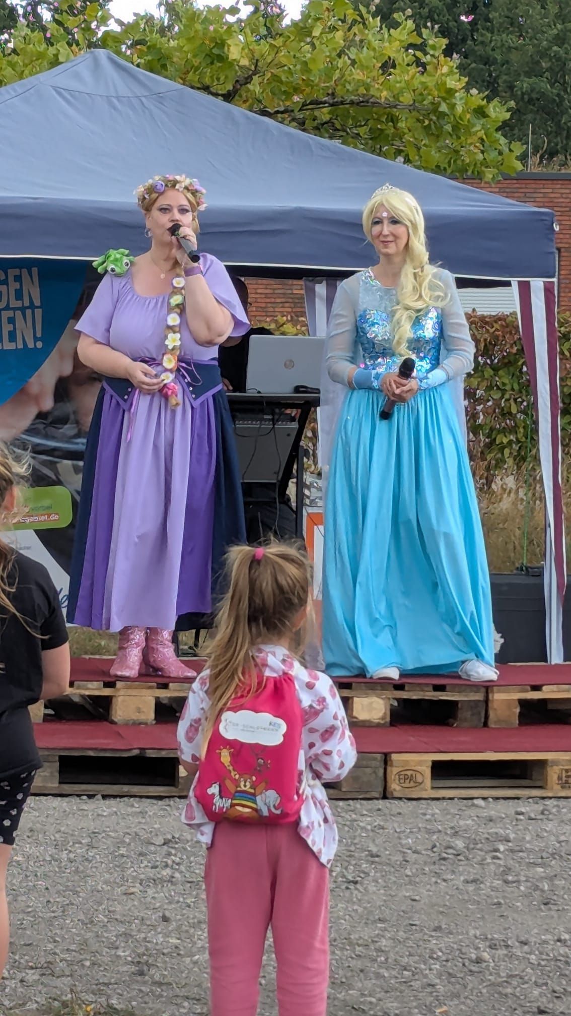 Zwei kostümierte Prinzessinnen auf der Bühne, einem Kind vor einem Veranstaltungszelt im Freien gegenüberstehend.