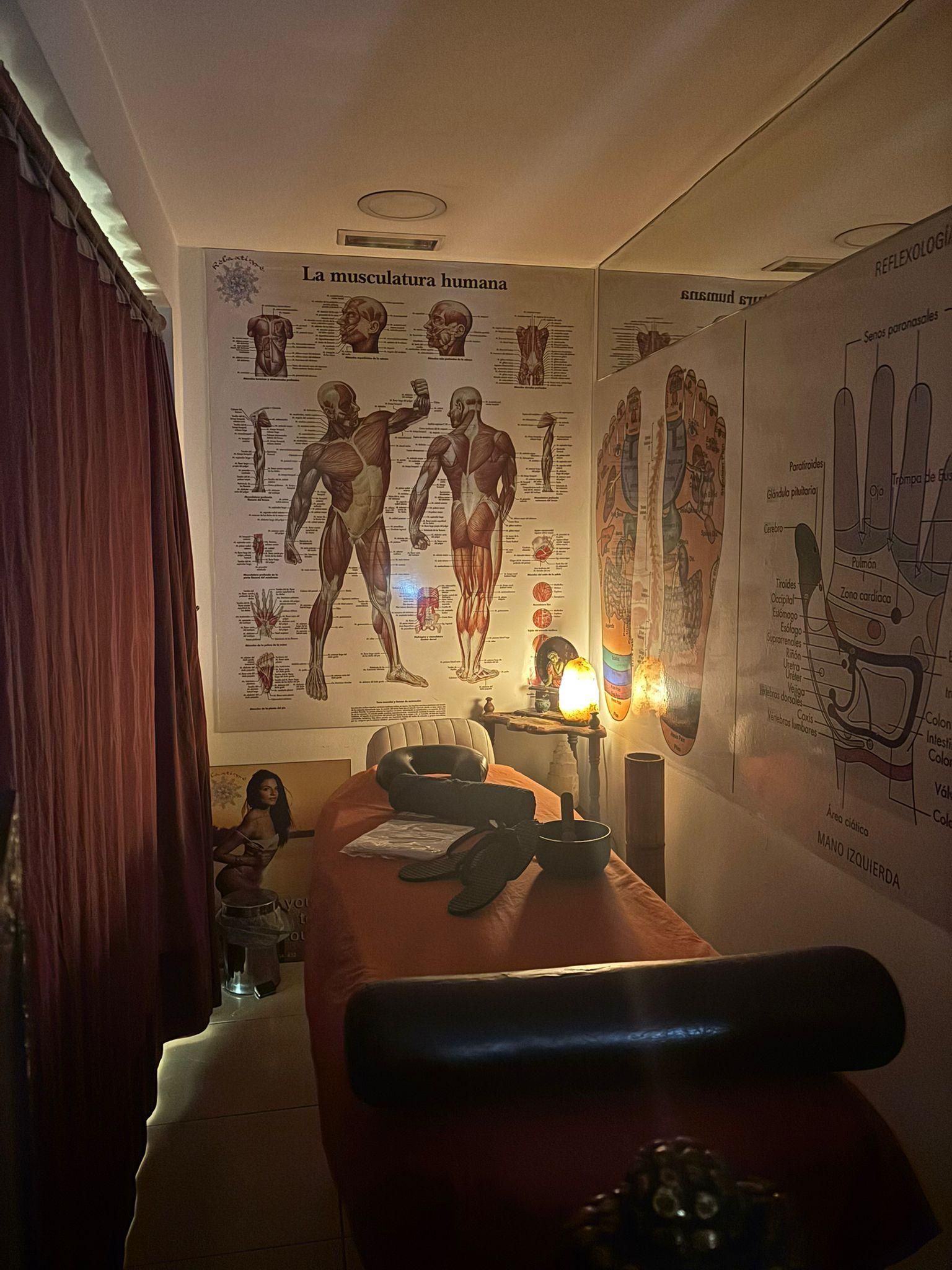 Sala de terapia de masajes con cuadros anatómicos en las paredes, mesa de masajes y cortinas.