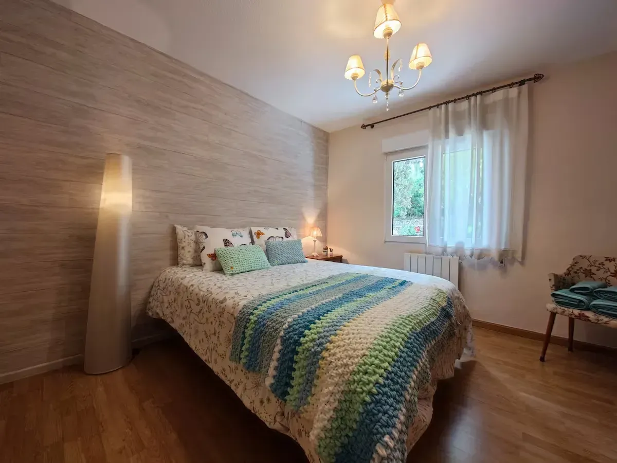 Dormitorio con cama, manta estampada, pared decorativa, lámpara, ventana y silla.