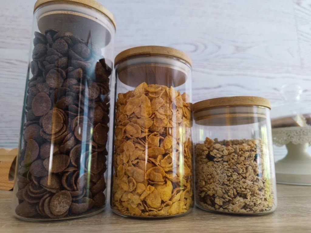 Tres frascos de vidrio llenos de diferentes cereales, tapas de madera, sobre un fondo de madera clara.