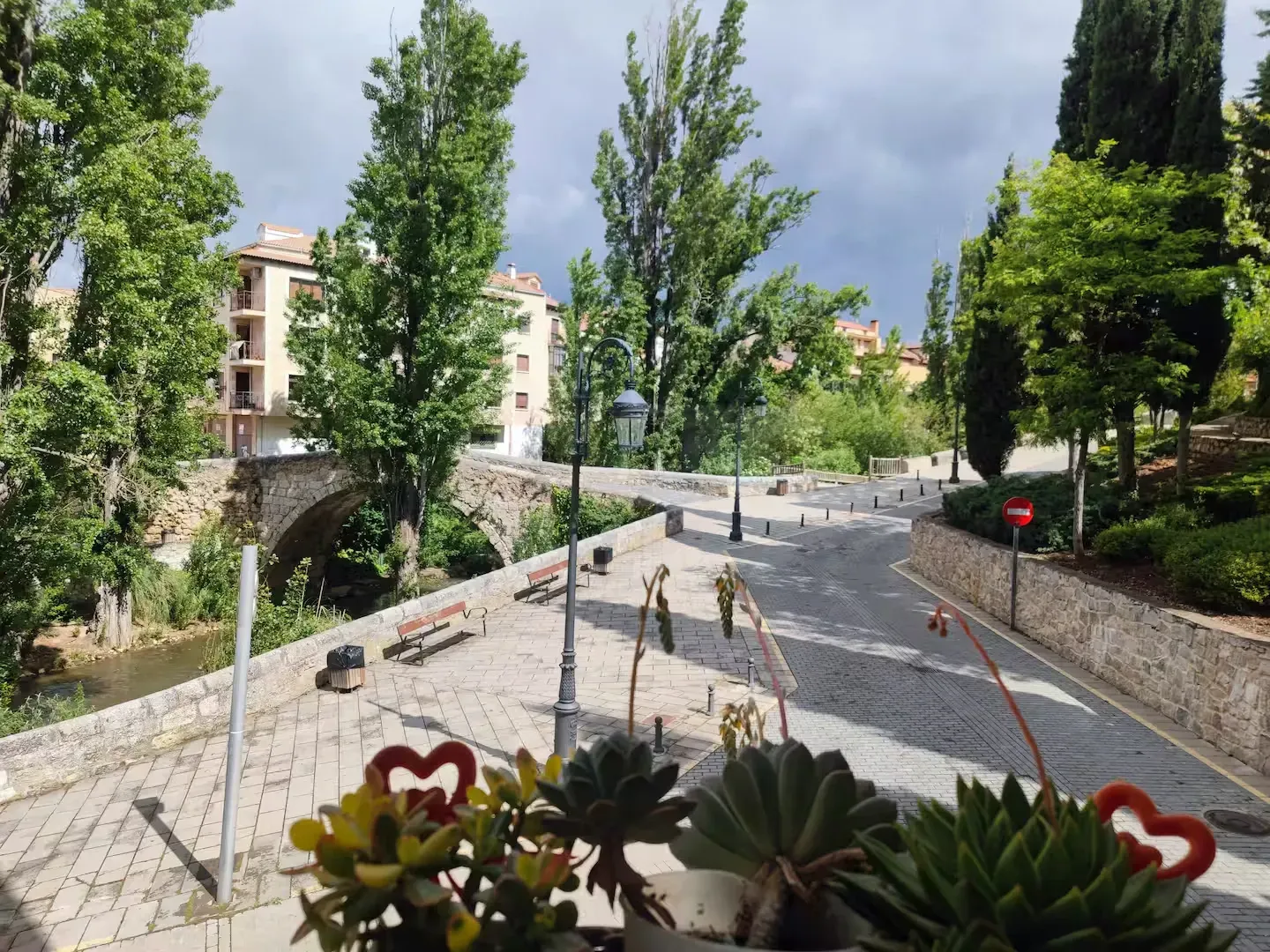 Puente de piedra sobre un río, con edificios y árboles. Un camino pavimentado se aleja del puente. Cielo nublado.