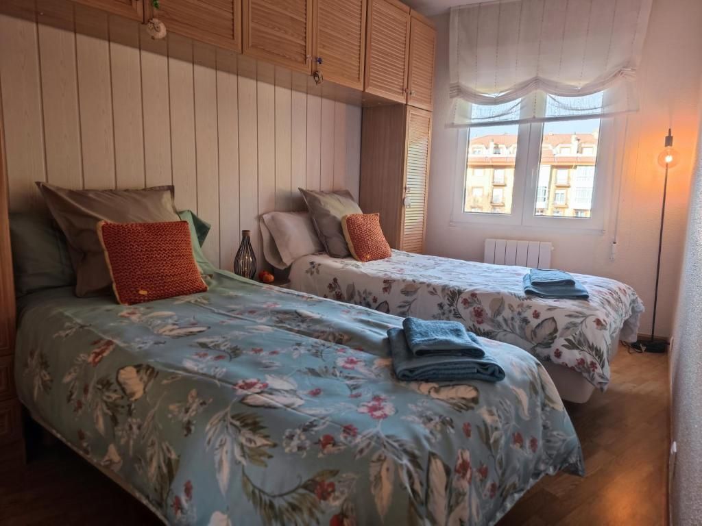 Dos camas individuales en una habitación con colchas florales, almohadas naranjas y una ventana con una cortina transparente.