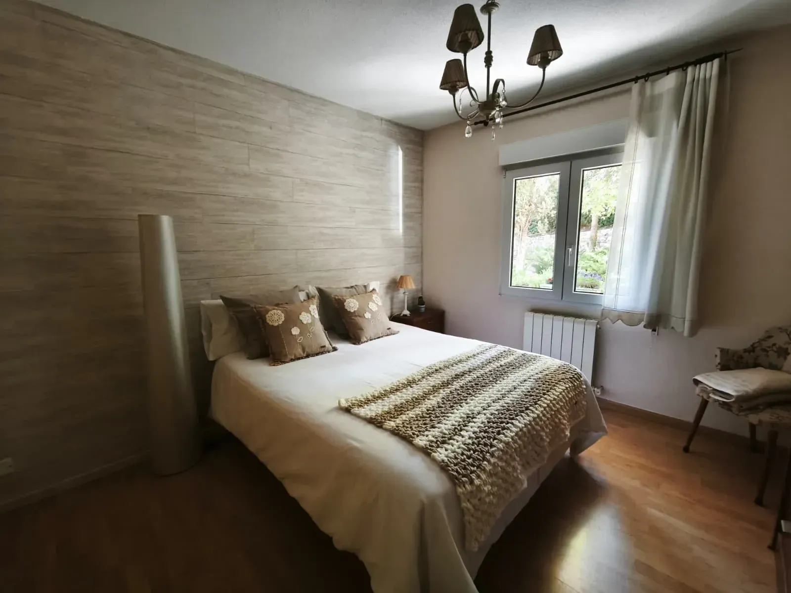 Dormitorio con cama, manta estampada, pared decorativa, lámpara, ventana y silla.