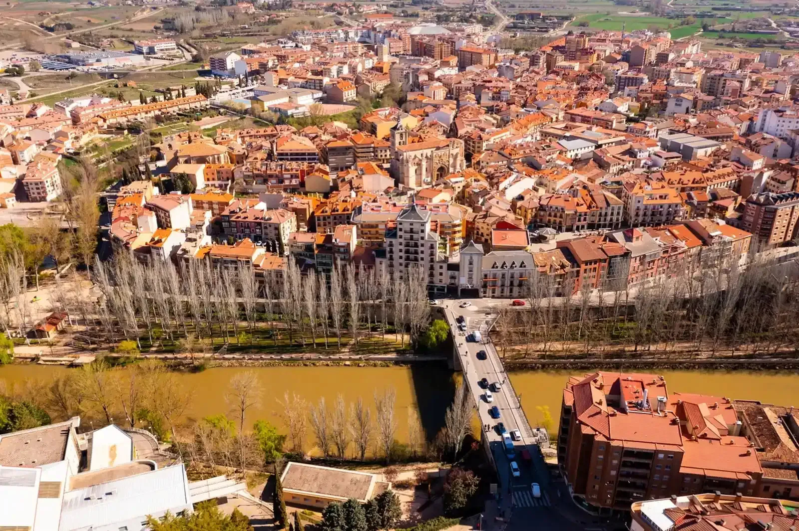 Vista aérea de una ciudad española con tejados de tejas rojas, un río y puentes. Día soleado con árboles y edificios.