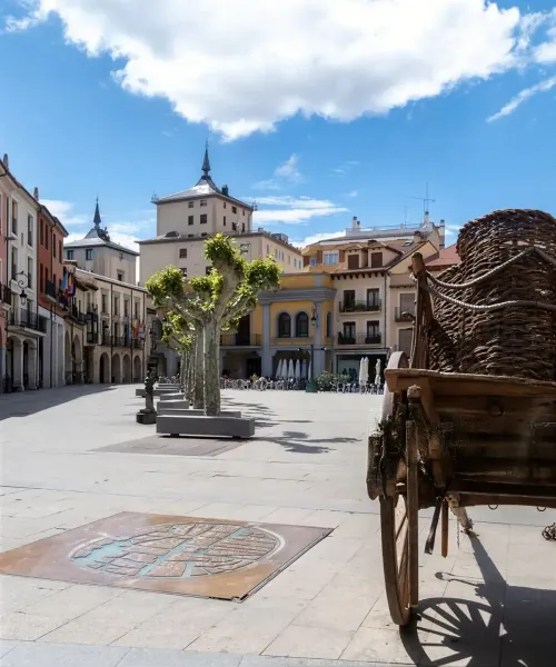 Plaza vacía en un pueblo europeo con edificios ornamentados, carros y árboles. Cielo azul con nubes.