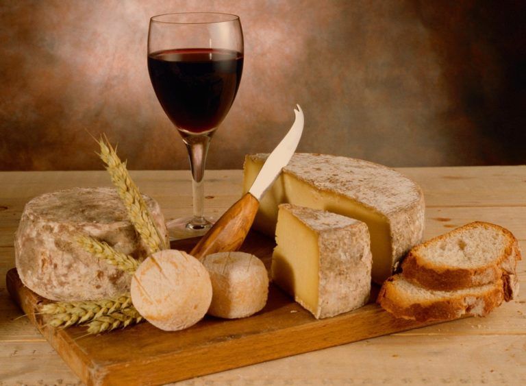 Copa de vino junto a varios quesos, pan y un cuchillo de queso sobre una tabla de madera.