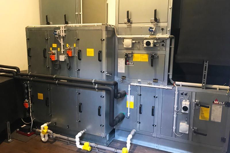 Große HLK-Einheit in einem Raum mit verschiedenen Rohren, Ventilen und Bedienfeldern. Grau und Schwarz mit gelben Akzenten.