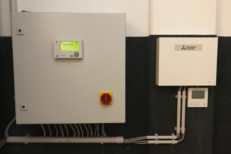 Zwei graue Schaltkästen an einer schwarz-weißen Wand, an deren Unterseite Kabel verlaufen.