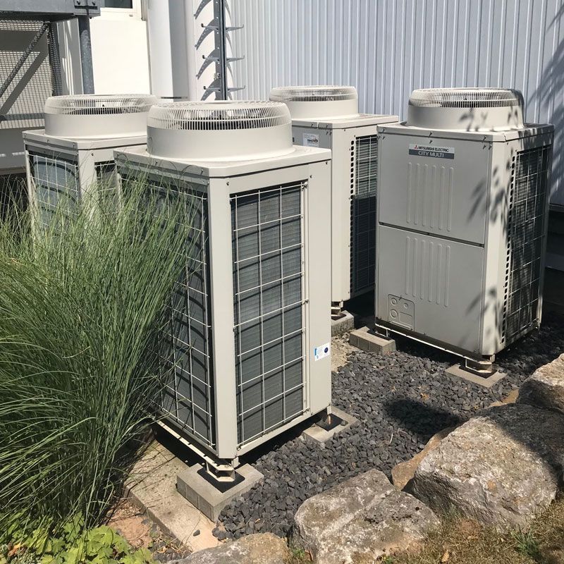 Mehrere Außen-HVAC-Einheiten auf Kies neben Büschen und Felsen.