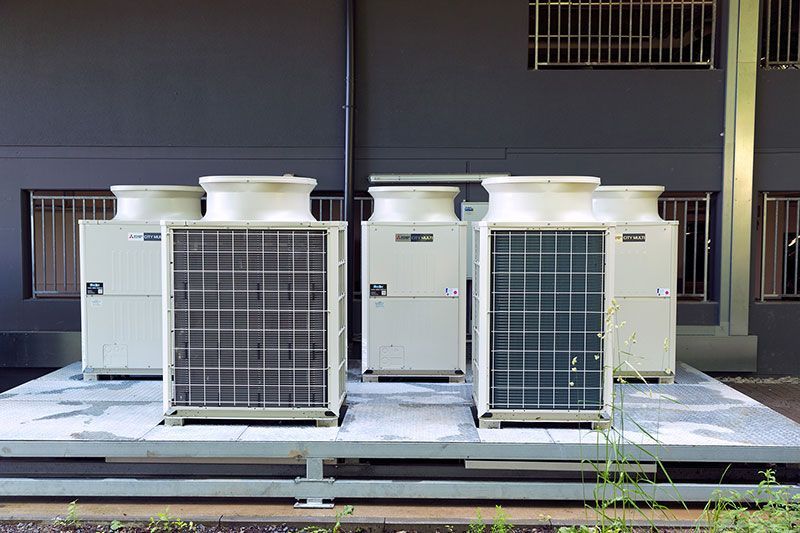 Fünf Klimaanlagen auf einer Plattform vor der dunklen Fassade eines Gebäudes.