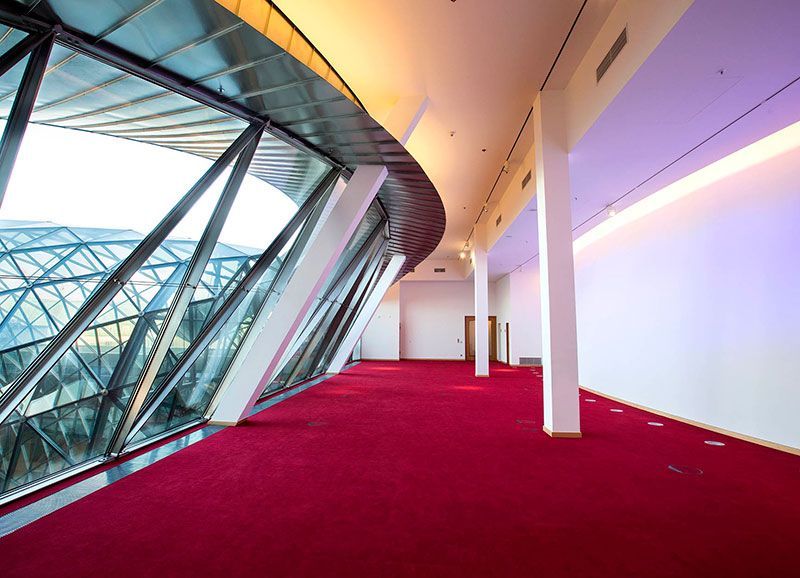 Innenansicht eines modernen Gebäudes mit rotem Teppich, weißen Säulen, großem schrägen Glasfenster und gewölbter Decke.