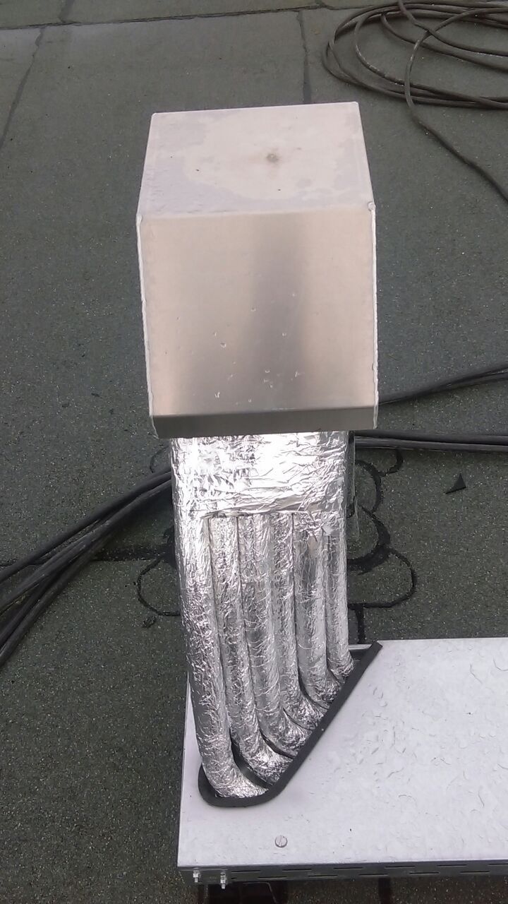 Silberner Metallkamin mit quadratischer Oberseite und mehreren gewellten Rohren auf einem Flachdach.