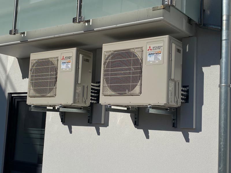 Zwei beige Klimaanlagen von Mitsubishi, an einer weißen Wand unter einer grauen Markise montiert.