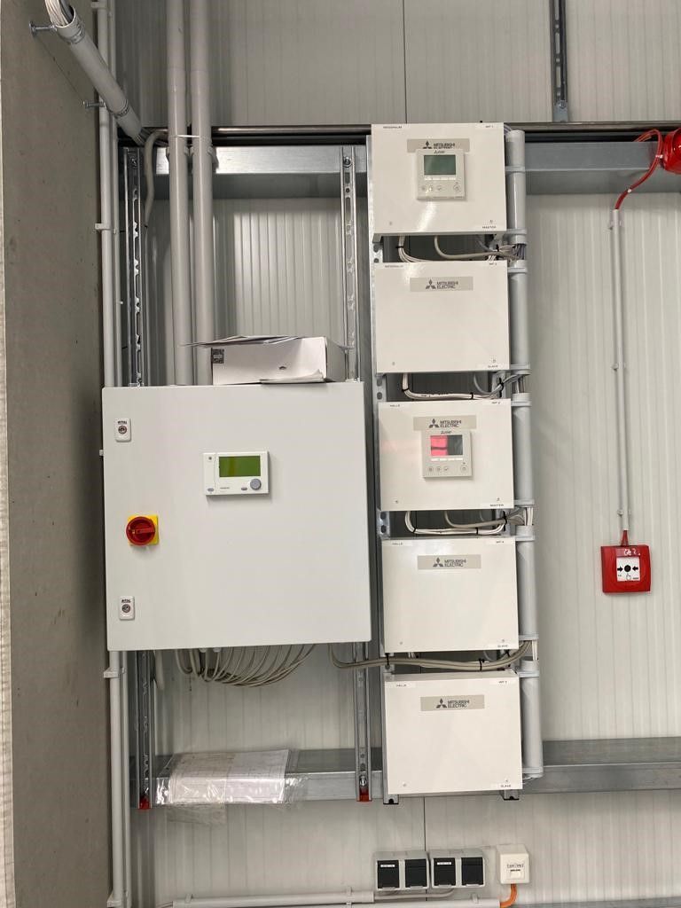 An einer Wand montierte Schalttafel und Wechselrichter, wobei verschiedene Messgeräte und Verkabelungen sichtbar sind.