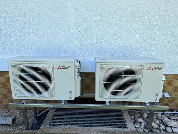 Zwei beigefarbene Klimaanlagen von Mitsubishi, montiert auf einem Metallständer vor einer weißen Wand.