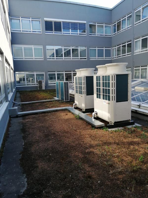 Zwei weiße Kühltürme auf einem braunen Dachgarten, umgeben von einem grauen Gebäude mit mehreren Fenstern.