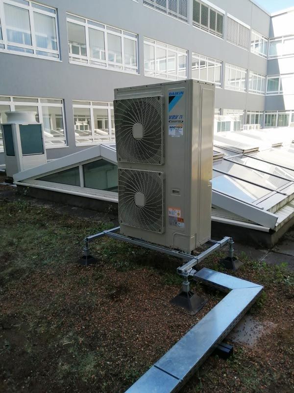 Klima- und Lüftungsanlage auf einem Dach. Beige, rechteckige Anlage mit zwei Ventilatoren auf einem Metallständer. Grünes Dach, Gebäude im Hintergrund.