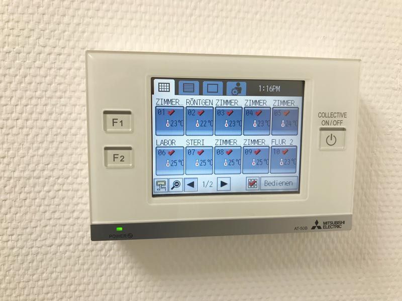 Weißer Mitsubishi HVAC-Regler mit einem Bildschirm, der Raumtemperaturen und Einstellungen anzeigt; an einer strukturierten weißen Wand montiert.