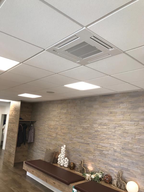 Wartezimmer mit Klimaanlage an der Decke, quadratischen Leuchten und einer Bank vor einer strukturierten Wand.