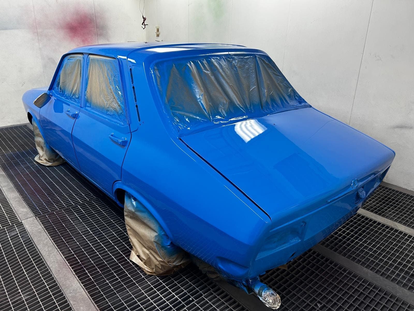 Coche azul en una cabina de pintura, con ventanas y ruedas cubiertas, mostrando pintura recién aplicada.