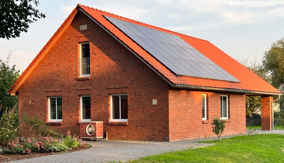 Ein Backsteinhaus mit rotem Ziegeldach und Solarpaneelen, eingebettet in einen Hof mit Garten und Auffahrt.