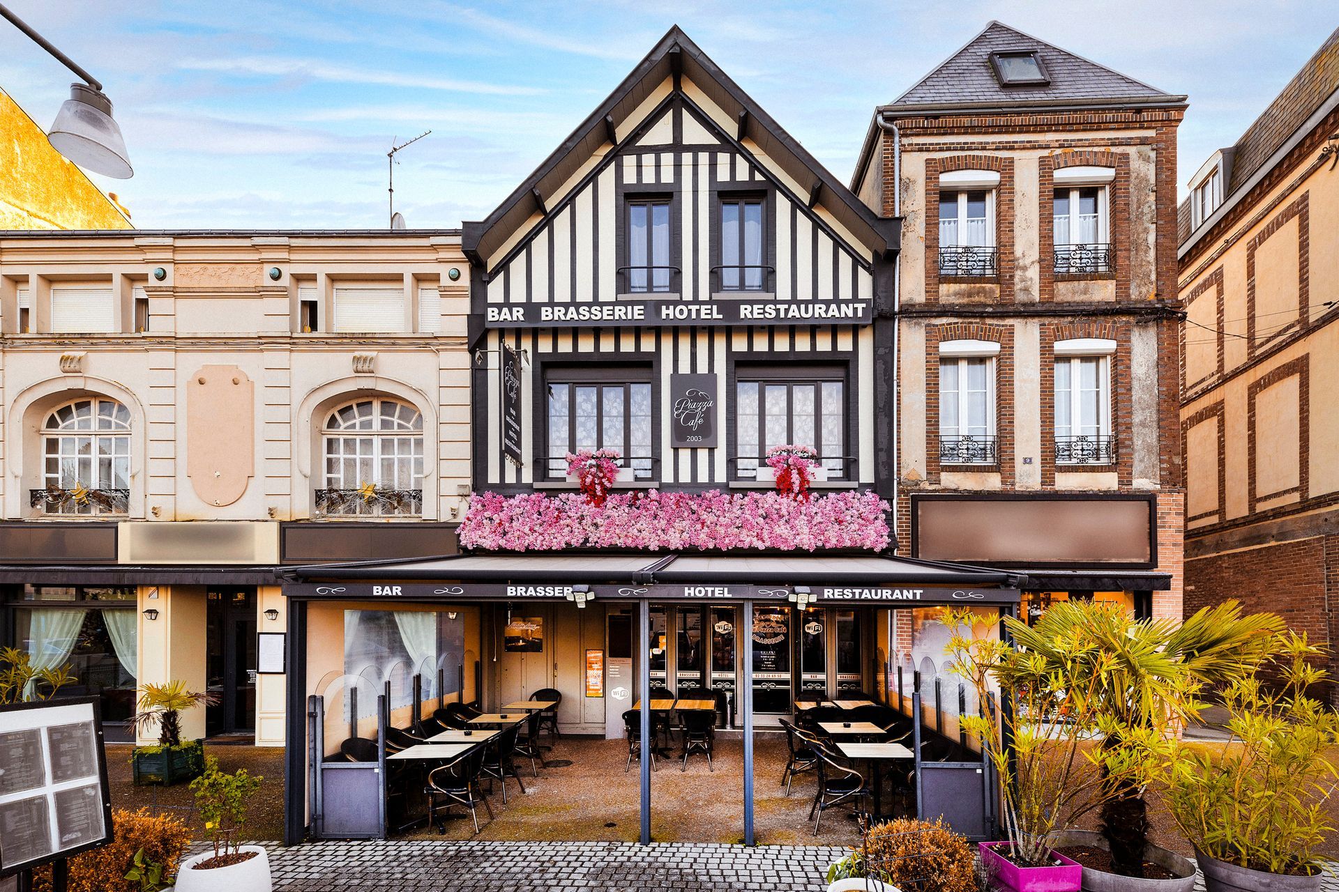 Devanture de l'HÔTEL BRASSERIE PIAZZA CAFÉ avec fleurs roses et auvent noir.