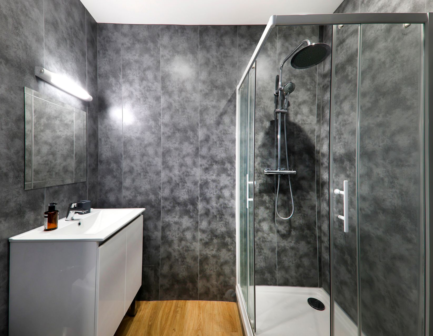 Salle de bains avec murs de carreaux gris texturés, douche en verre, meuble avec lavabo blanc et parquet.