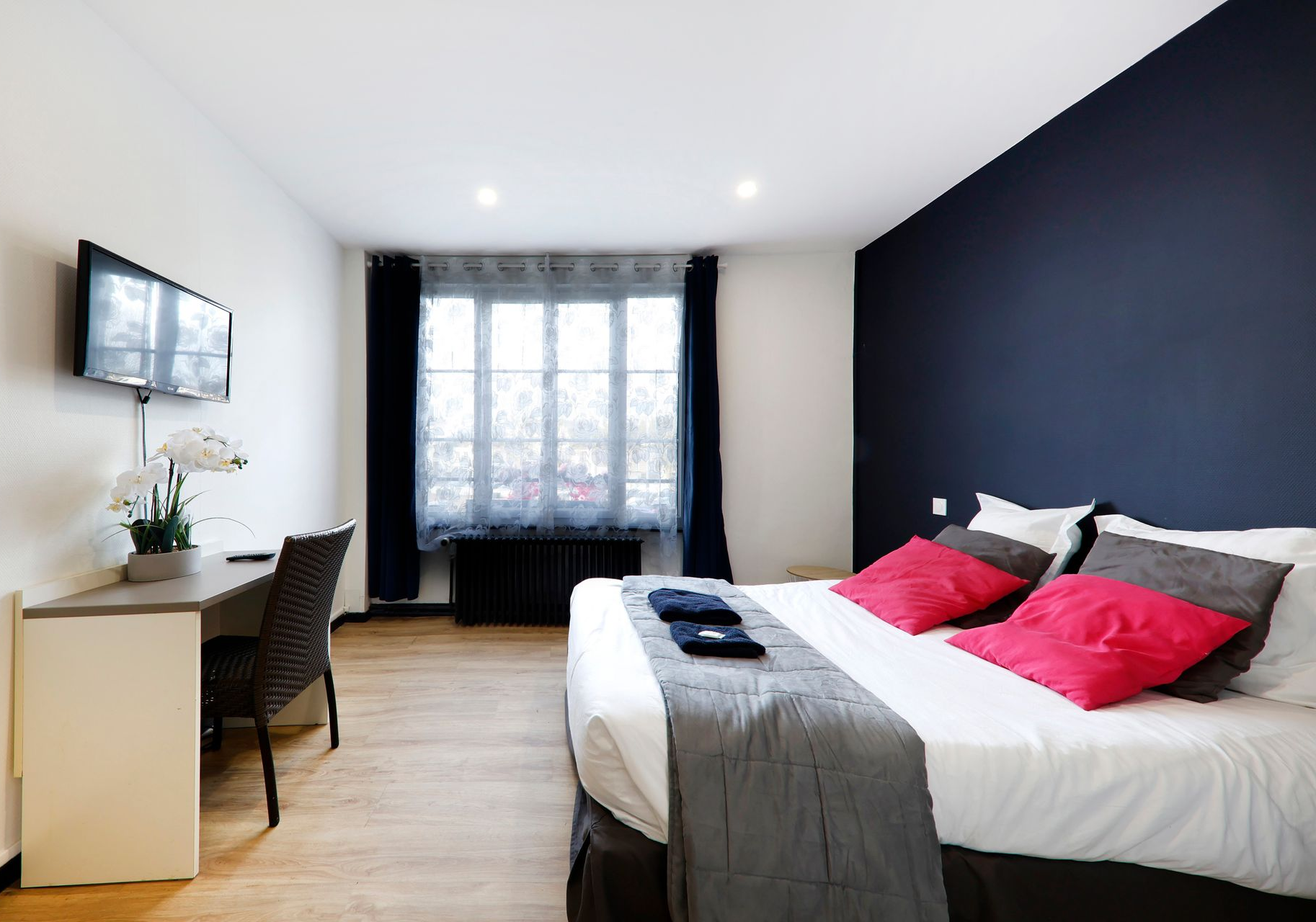 Chambre avec lit, bureau, télévision et fenêtre ; mur aux accents bleu marine, coussins roses et gris.