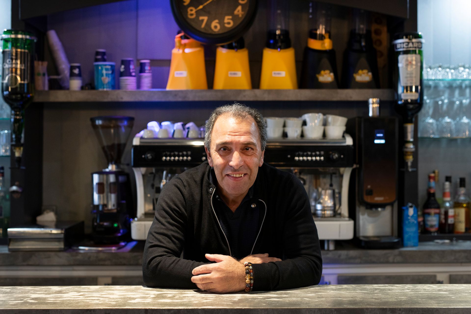 Le patron de l'HÔTEL BRASSERIE PIAZZA CAFÉ derrière le bar, les bras croisés. On aperçoit en arrière-plan des machines à café, des bouteilles.
