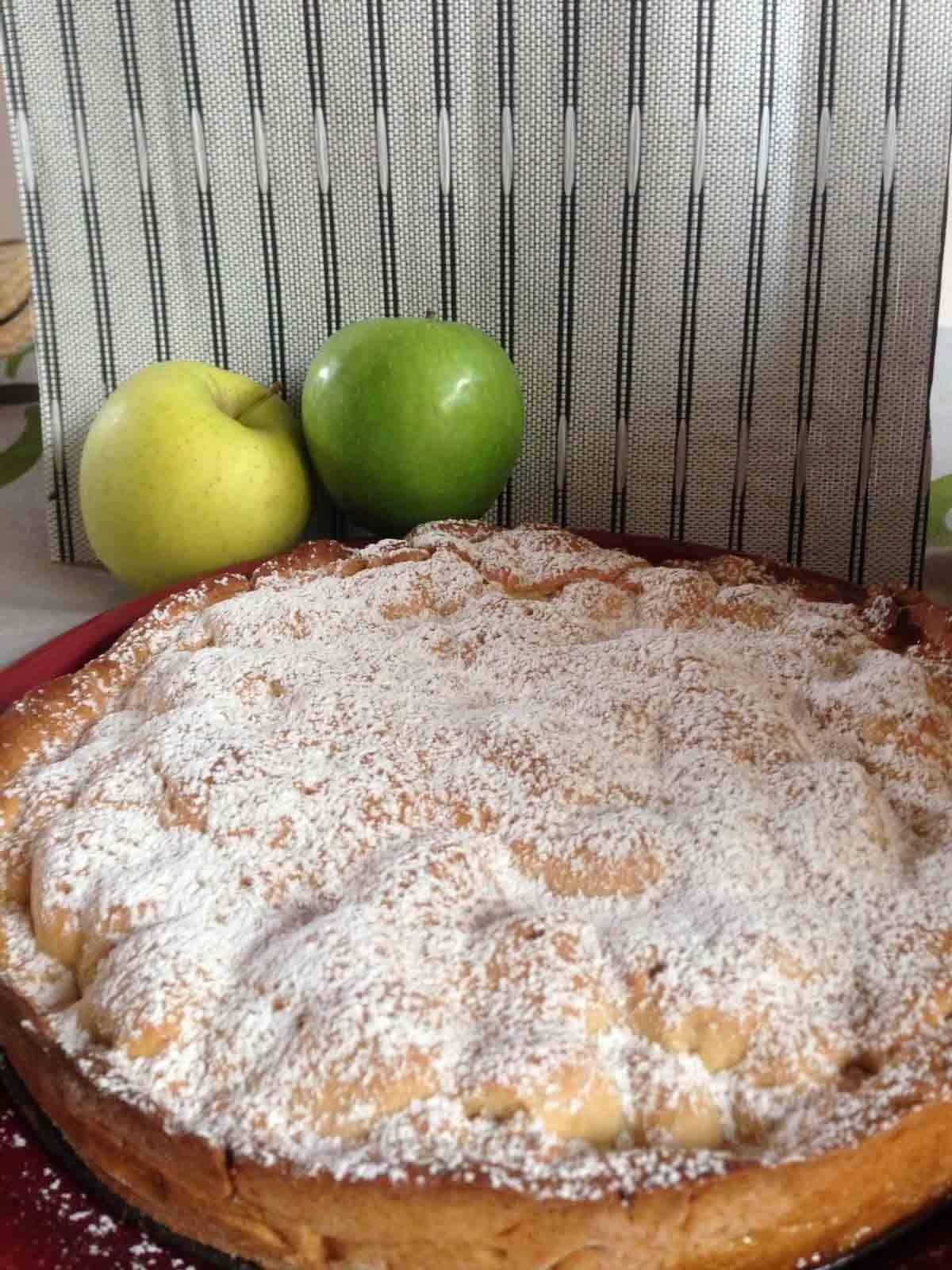 Un pastel con azúcar en polvo encima está sobre una mesa al lado de manzanas.