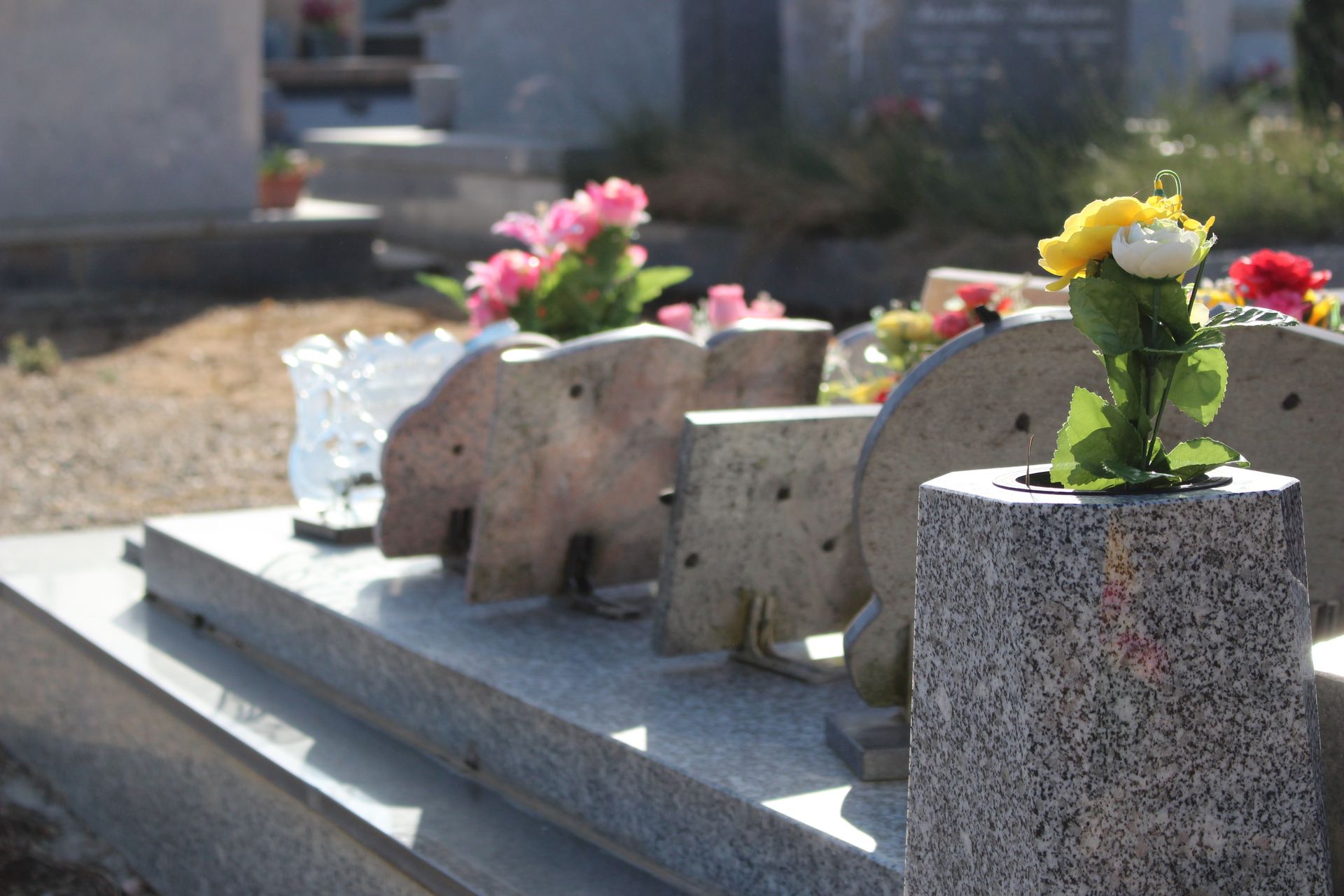 Des pierres tombales ornées de compositions florales dans une allée ensoleillée d'un cimetière.