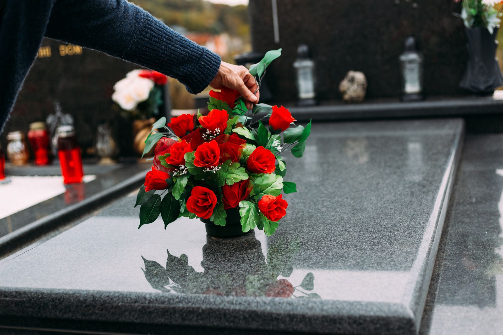 Une main dépose des roses rouges sur une pierre tombale polie dans un cimetière.