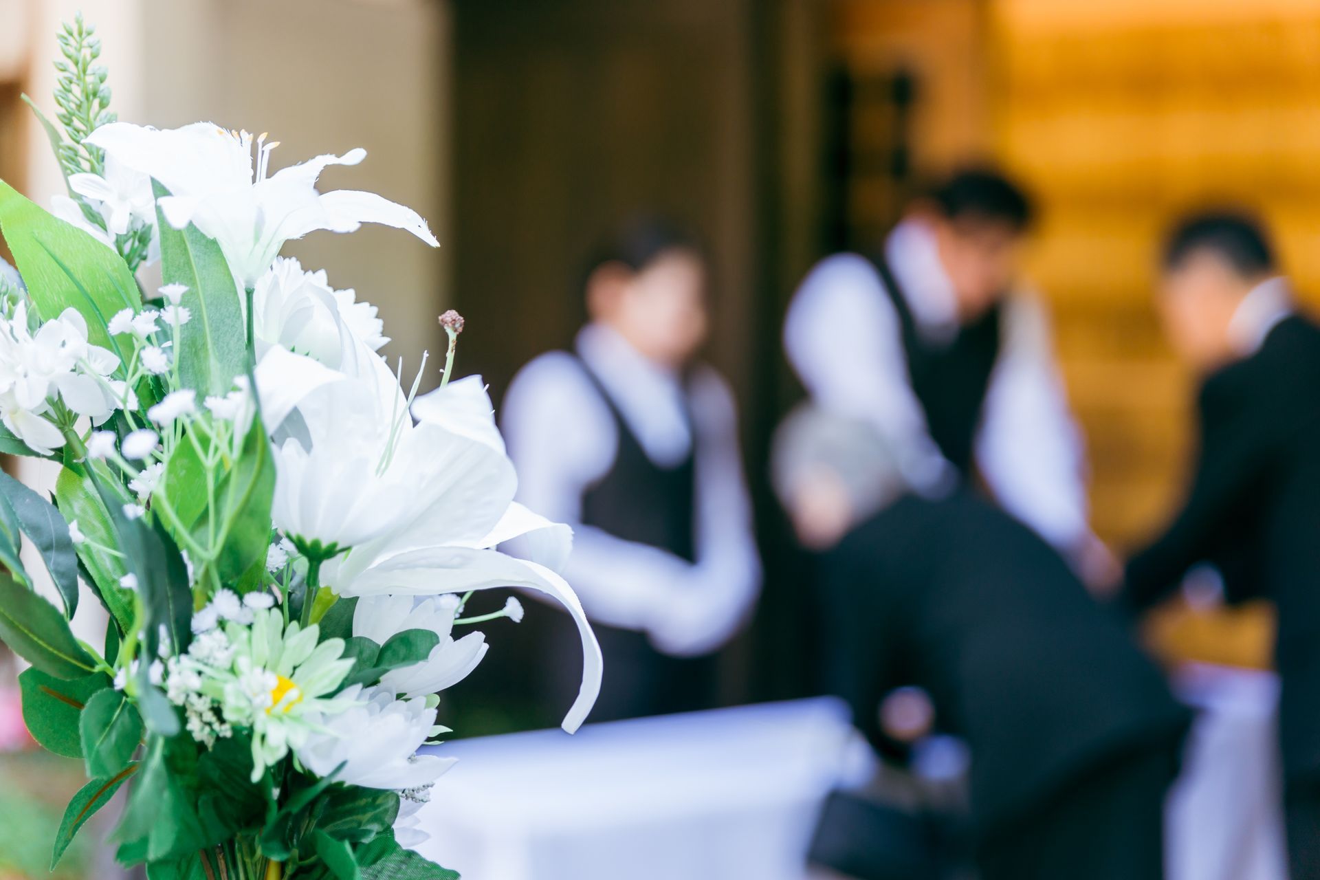 Composition florale blanche nette, avec des invités flous en tenue de soirée lors d'une réception de mariage.