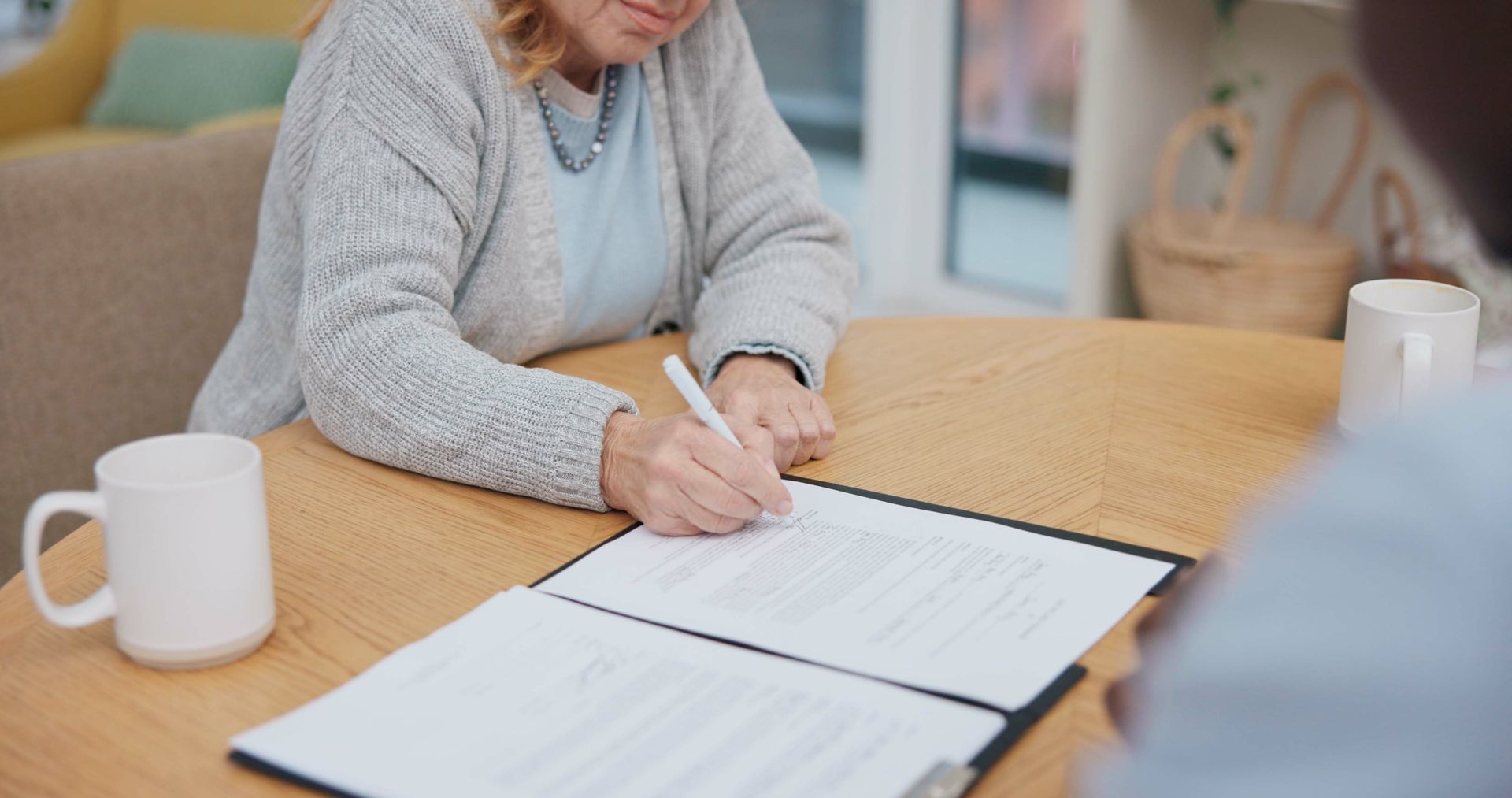 Une personne signe un document à une table en bois, une tasse à proximité.