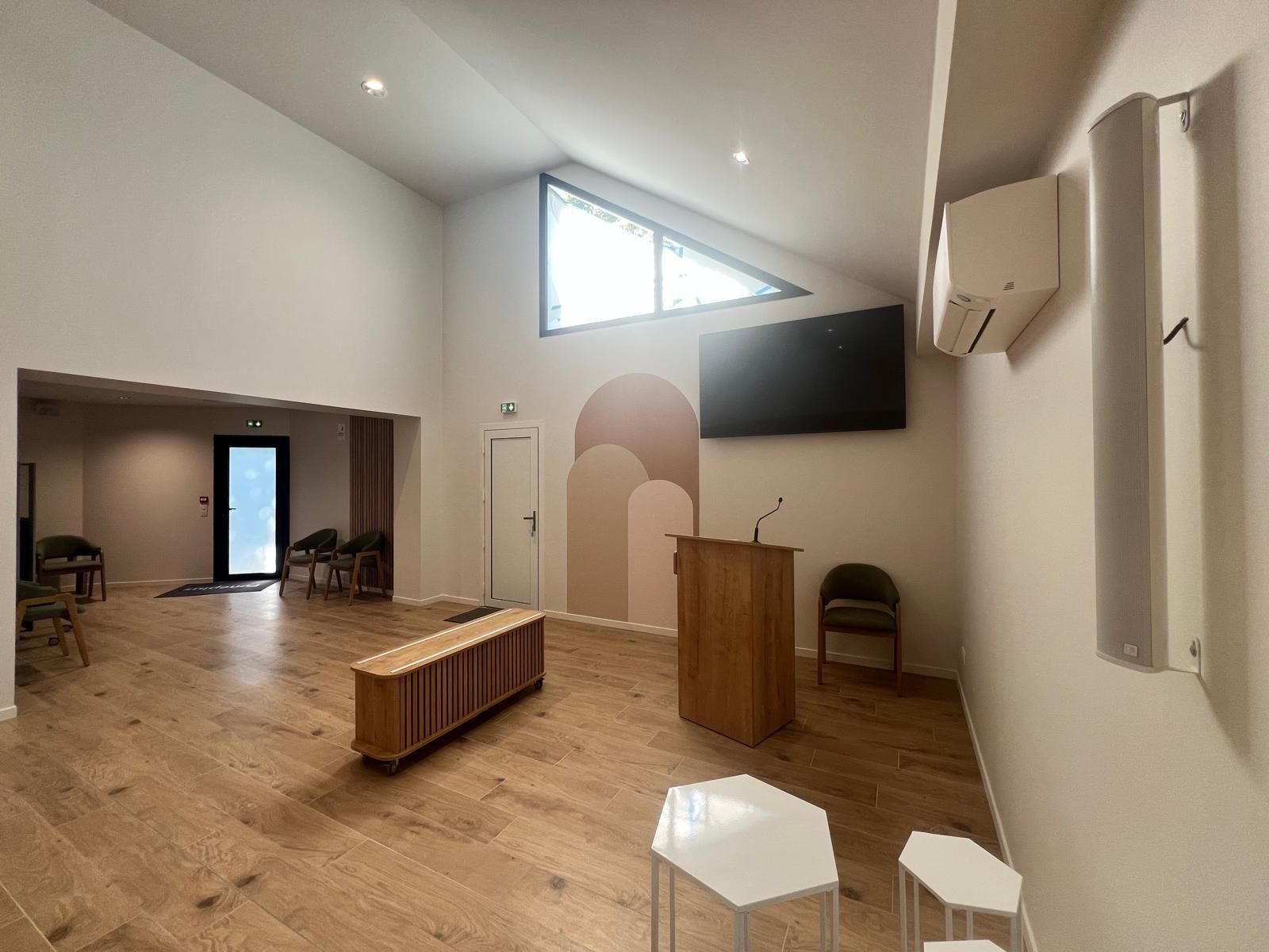 Salle moderne et spacieuse avec de hautes fenêtres, un banc en bois, une télévision, des chaises et un pupitre sur un parquet clair.