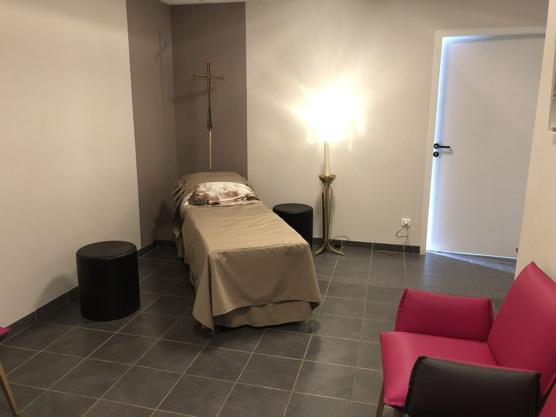 Chambre funéraire avec un lit, des tabourets noirs et une chaise rose sur un sol carrelé.